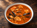 A richly colored seafood stew with fish and shellfish, warm lighting, and a rustic wood setting. Signed Dr. E World Culture and Education.